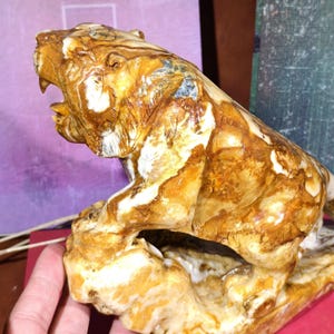 May include: A detailed sculpture of a tiger in a roaring pose, carved from a stone with brown and white marbling. The tiger is depicted with its mouth open, revealing its teeth, and is mounted on a naturalistic base.