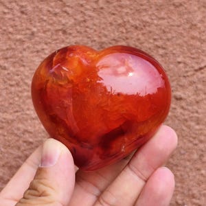 May include: A polished, heart-shaped carnelian gemstone. The stone is a vibrant mix of red and orange hues, with a glossy, reflective surface. The gemstone is held in a hand, showcasing its smooth, rounded form.