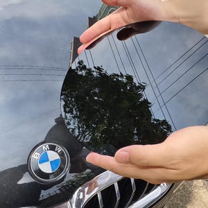 May include: A black, circular object is held above a black car hood with a BMW emblem. The object reflects the sky, trees, and power lines. The car hood is black and shiny.