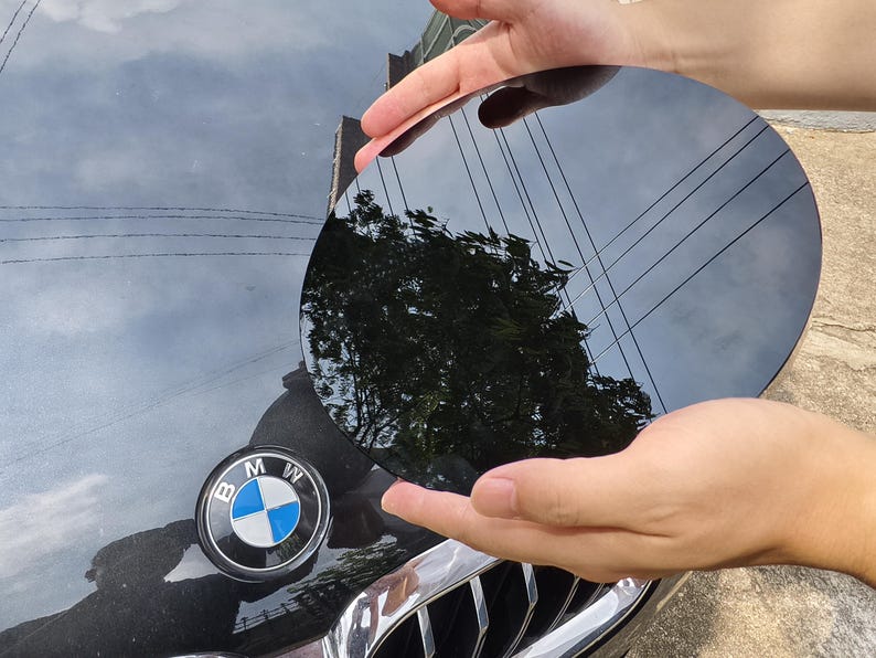 May include: A black, circular object is held above a black car hood with a BMW emblem. The object reflects the sky, trees, and power lines. The car hood is black and shiny.