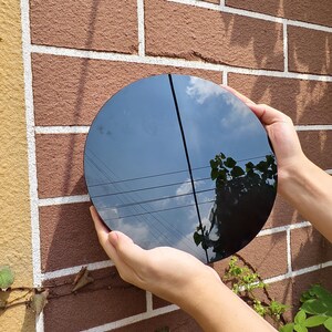 May include: A round, black mirror reflecting a blue sky with clouds, power lines, and tree branches. The mirror is held against a red brick wall, showcasing its reflective surface and circular shape.