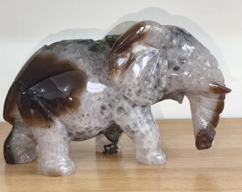 May include: A carved elephant figurine made of translucent, crystalline stone with a polished finish. The sculpture features a mottled pattern of light grey, white, and deep brown patches throughout its body, ears, and trunk, showing natural mineral inclusions.