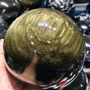 Könnte beinhalten: Eine polierte Gold-Obsidian-Kugel mit einer glatten, reflektierenden Oberfläche und wirbelnden Einschlüssen in Metallic-Gold und Tiefschwarz. Der Stein weist einen natürlichen Glanz auf, der das Licht einfängt.