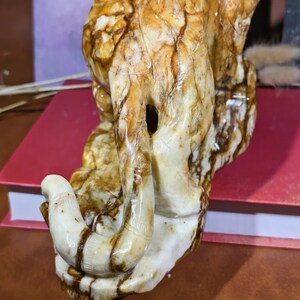 May include: A carved stone sculpture of an elephant, featuring a curved trunk and detailed legs. The sculpture is primarily cream-colored with brown veining, giving it a natural, earthy appearance. The elephant's trunk is curved and detailed.