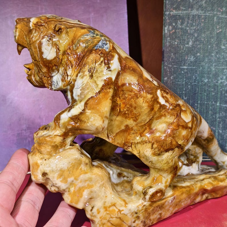 May include: A detailed sculpture of a tiger in a roaring pose, carved from stone. The tiger's fur is depicted in shades of brown, tan, and white, with visible texture. The sculpture is mounted on a base that mimics a natural rock formation.