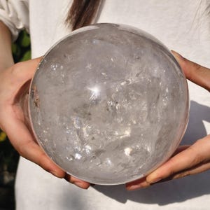 May include: A large, spherical clear quartz crystal ball with a smooth, polished surface. The interior of the sphere displays natural inclusions, fractures, and internal reflective facets that create a complex, textured appearance throughout.