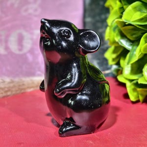 Pode incluir: Uma estatueta de rato preto brilhante com um design detalhado. O rato está em pé, com a cabeça inclinada para cima. A estatueta tem orelhas, patas e cauda visíveis. O fundo é uma mistura desfocada de vermelho e verde.