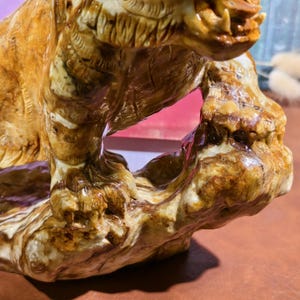May include: A detailed, close-up view of a carved sculpture of a tiger. The sculpture is primarily shades of brown and tan, with intricate details. The tiger's mouth is open, revealing teeth. The sculpture is mounted on a base.