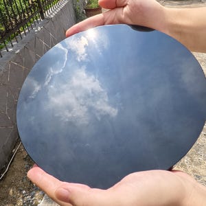 May include: A round, dark gray object reflecting a cloudy sky. The object is held by two hands, with the sky's blue and white clouds mirrored on its surface. The object appears to be smooth and polished.