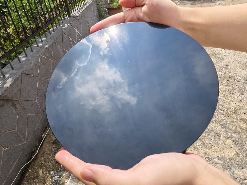 May include: A round, dark gray object reflecting a cloudy sky. The object is held by two hands, with the sky's blue and white clouds mirrored on its surface. The object appears to be smooth and polished.