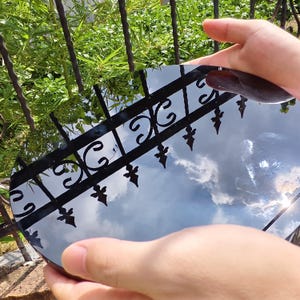 May include: A round, reflective surface held by hands, reflecting a cloudy sky and a black wrought iron fence. The surface appears to be a mirror or polished material, with a dark brown object on top.