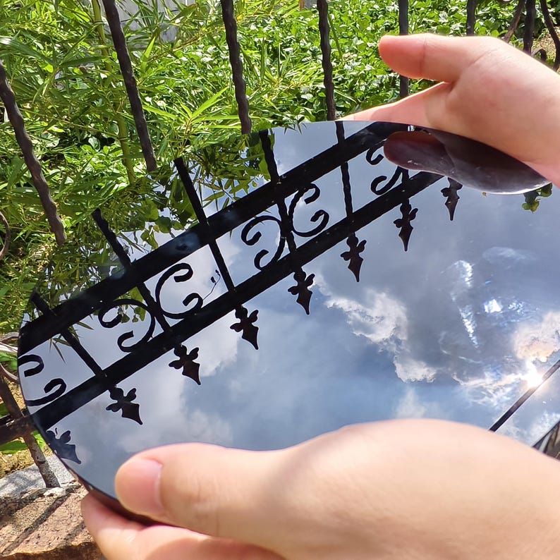 May include: A round, reflective surface held by hands, reflecting a cloudy sky and a black wrought iron fence. The surface appears to be a mirror or polished material, with a dark brown object on top.