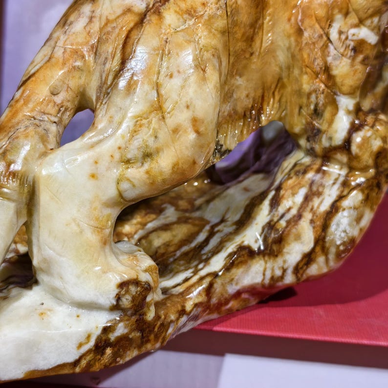 May include: Close-up of a carved sculpture with a cream and brown marbled pattern. The sculpture has a smooth, polished surface with a naturalistic design. The details of the sculpture are visible, showcasing the craftsmanship.