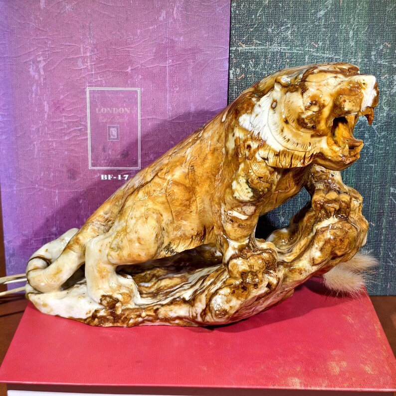 May include: A detailed sculpture of a roaring tiger, carved from a light brown material with darker brown and white accents. The tiger is posed on a textured base, with a small, fluffy tail. The background includes a purple book with the text "LONDON" and "BF-17".