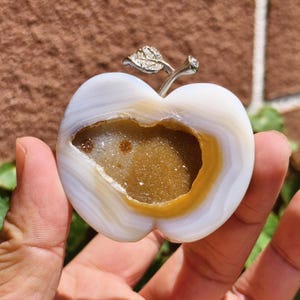 May include: A white and tan agate apple sculpture with a silver stem and leaf. The apple has a hollowed-out centre filled with sparkling golden crystals. The sculpture is held in a hand.