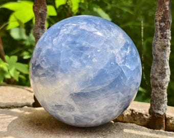 May include: A large, spherical blue calcite crystal ball. The crystal has a cloudy, translucent appearance with varying shades of blue and white. The sphere is resting on a stone surface, with green foliage in the background.