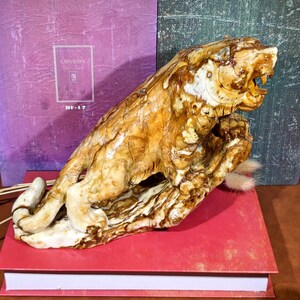 May include: A detailed sculpture of a tiger in a roaring pose, carved from a light brown material with darker brown accents. The tiger rests on a base, with a small tuft of fur visible. The sculpture is placed on a red book.