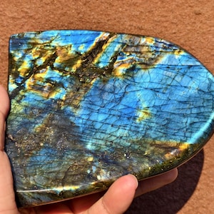 May include: A polished labradorite stone displaying blue, gold, and green iridescence. The gemstone is held in a hand, highlighting its unique patterns and textures. The stone has a smooth, curved shape.