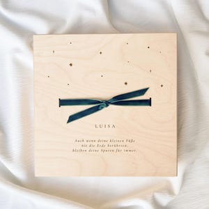 Könnte beinhalten: Helle Holzplakette mit dem Namen "LUISA" und einem tealfarbenen Band. Der Hintergrund ist helles Holz mit Sternen. Der Text auf Deutsch lautet: "Auch wenn deine kleinen Füße nie die Erde berühren, bleiben deine Spuren für immer."