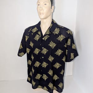 May include: Black short-sleeved shirt with a gold floral pattern. The shirt has a classic collar and a single chest pocket. The shirt is displayed on a mannequin.