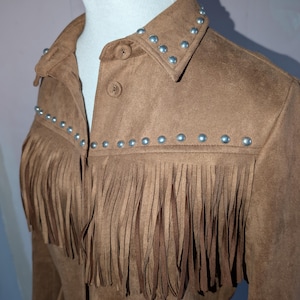 May include: A brown suede jacket with fringe and silver stud embellishments. The jacket has a collared neckline and button closure. Fringing is featured on the chest and sleeves, giving a western style.