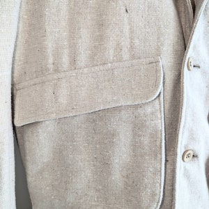 May include: A light beige linen blazer with a large patch pocket on the left side. The blazer has a single button closure and a textured fabric.