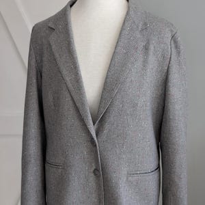 Op de afbeelding: Een grijze blazer met een gespikkeld patroon, weergegeven op een mannequin. De jas heeft een sluiting met twee knopen, een revers en twee voorzakken. De stof heeft vlekjes rood en wit.