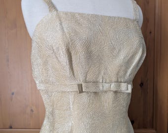 May include: A gold-colored, sleeveless dress with a square neckline and thin shoulder straps. The dress features a subtle, textured pattern and a small bow detail at the waist. The dress is displayed on a mannequin.