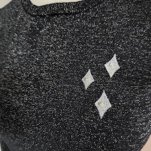 Può includere: Top in maglia nera con fili metallici argentati. Il top presenta tre abbellimenti bianchi a forma di diamante, ciascuno con un piccolo centro a forma di perla. Lo scollo e i polsini sono a coste.