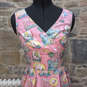 Op de afbeelding: Een roze jurk zonder mouwen met een V-hals en een flamingo print. De jurk heeft een getailleerd bovenstuk en een volle rok. De print bevat roze flamingo's, palmbomen en de woorden "Sunset" en "Flamingos".