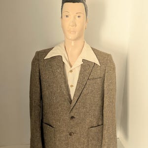 May include: A brown tweed blazer with a textured weave, featuring two front patch pockets, a notched lapel, and two dark buttons, worn over a cream-coloured button-down shirt with a wide collar on a mannequin with a dark pompadour hairstyle.