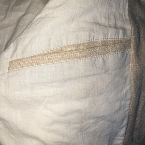 May include: Close-up of the inside of a garment, showing a beige linen fabric with a seam detail. The seam is made of a woven beige fabric and is stitched with a straight stitch.