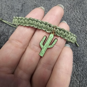 May include: A green macrame bracelet featuring a light green cactus charm with black outlines and gold-colored accents. The bracelet is made of woven green cord and is displayed on a gray surface.