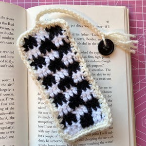 May include: A handmade crochet bookmark with a black and white houndstooth pattern. The bookmark has a cream-colored border and a loop with a black button and fringe. The bookmark is placed on an open book.