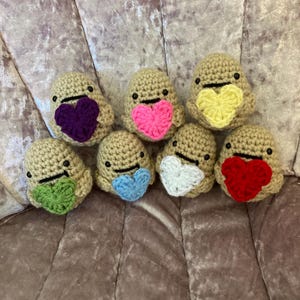May include: A collection of seven beige crochet figures, each with a heart-shaped accent in various colors: purple, pink, yellow, green, blue, white, and red. The figures have small black eyes and a stitched smile. The items are arranged on a textured, light purple surface.