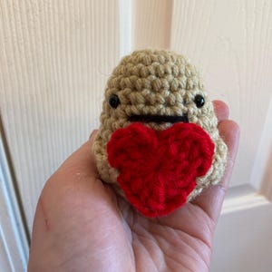 Pode incluir: Um brinquedo de pelúcia de crochê em forma de uma pequena figura com um coração vermelho. A figura é bege com olhos pretos e uma boca costurada preta. O coração é de cor vermelha vibrante.