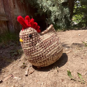 May include: A crocheted chicken toy in brown and beige tones, featuring a red comb and yellow beak. The handmade amigurumi chicken is positioned on the ground, set against a natural outdoor backdrop.