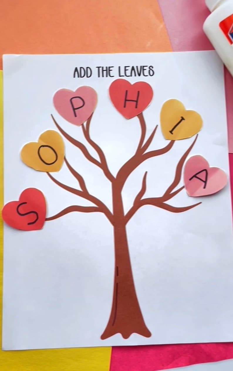 May include: A handmade craft project featuring a brown tree with heart-shaped leaves spelling "SOPHIA". The leaves are in shades of pink, yellow, and red. The phrase "ADD THE LEAVES" is written at the top. The background includes pink and orange paper.