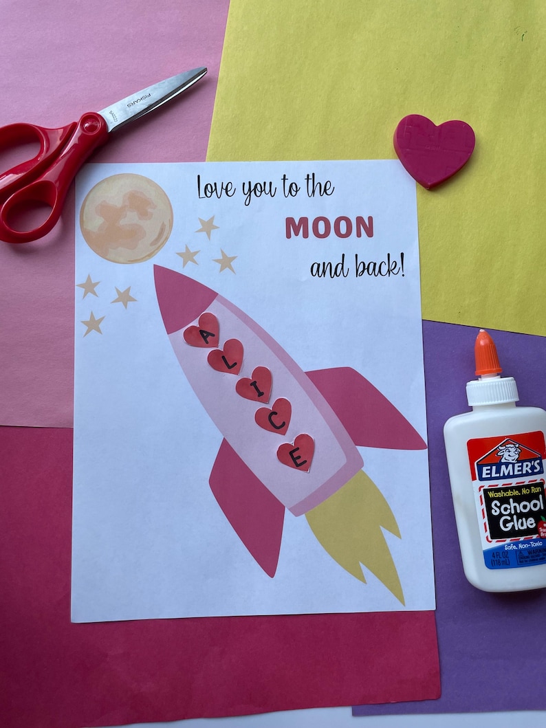 May include: A handmade card with a pink rocket ship design, the name "ALICE" in hearts, and the text "Love you to the MOON and back!". Includes scissors, glue, and a pink heart on a colorful background.