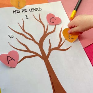 May include: A craft activity featuring a brown tree illustration with heart-shaped leaves. The text "ADD THE LEAVES" is above the tree. A child's hand is placing a heart-shaped leaf with the letter "E" on the tree. Markers and colorful paper are in the background.