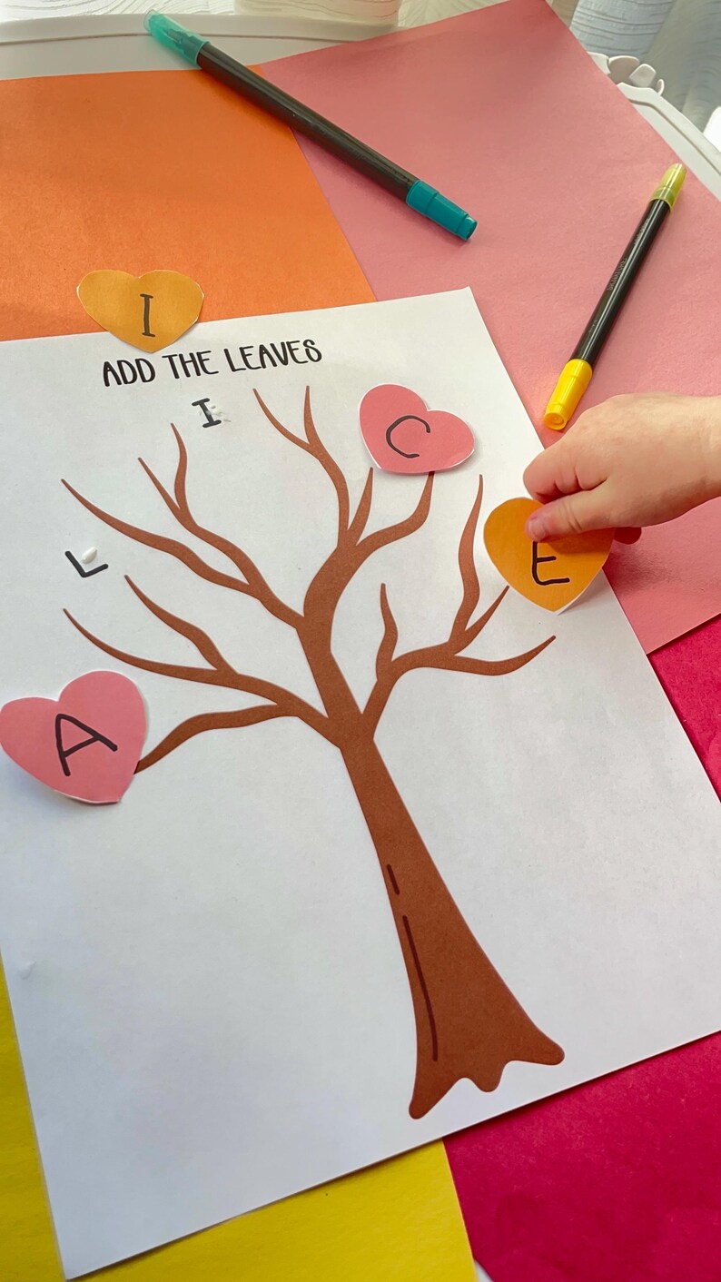 May include: A craft activity featuring a brown tree illustration with heart-shaped leaves. The text "ADD THE LEAVES" is above the tree. A child's hand is placing a heart-shaped leaf with the letter "E" on the tree. Markers and colorful paper are in the background.