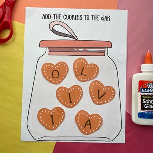 May include: A craft project featuring a drawing of a jar with heart-shaped cookies inside, spelling out the name "Olivia". The text "ADD THE COOKIES TO THE JAR" is above the jar. Scissors and glue are visible on the colorful background.