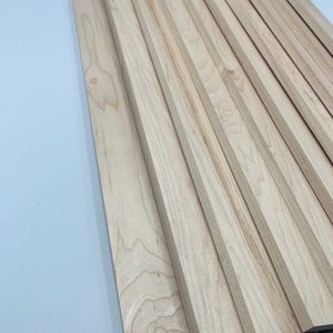Könnte beinhalten: Eine Sammlung unfertiger Holzbretter, diagonal angeordnet. Die Bretter haben eine helle, natürliche Holzfarbe mit sichtbaren Maserungsmustern. Die Kanten sind sauber und gerade, was darauf hindeutet, dass sie für ein Holzbearbeitungsprojekt oder eine Heimwerkerarbeit bereit sind.