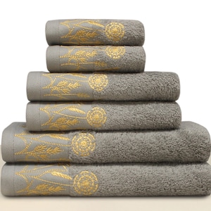 May include: A stack of six gray bath towels with gold embroidered floral designs. The towels are arranged in a pyramid shape, with the top towels smaller than the bottom ones. The embroidery includes a circular design and leaf-like patterns.