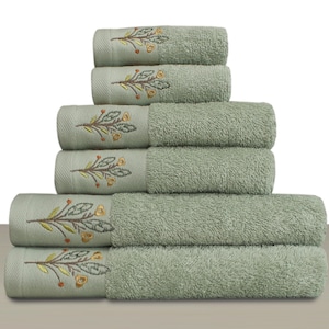 May include: A stack of six sage green bath towels. Each towel features an embroidered floral design in yellow, orange, and green. The towels are neatly rolled and stacked, showcasing a soft, textured appearance. The background is plain white.