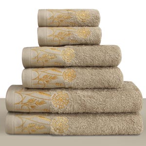 May include: A stack of six beige bath towels. Each towel features an embroidered gold floral design on the border. The towels are stacked neatly, showcasing the detailed embroidery and soft texture. Ideal for bathroom decor.