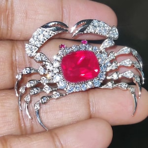 Pode incluir: Broche de caranguejo prateado com uma grande gema vermelha quadrada no centro. As garras e pernas do caranguejo são detalhadas com cristais transparentes e brilhantes. Uma pequena gema vermelha redonda está colocada acima.
