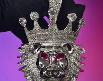May include: A silver-toned lion head pendant featuring a crown adorned with numerous small clear stones. The lion has a polished metallic finish, a detailed mane encrusted with clear stones, and two prominent red stones set as eyes.