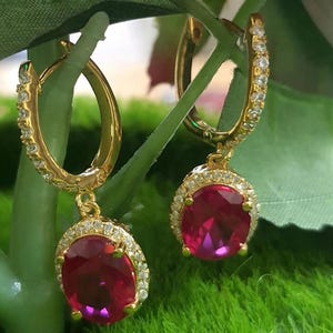 May include: A pair of gold-colored hoop earrings with a dangling, oval-shaped, red gemstone. The hoops are encrusted with small, clear stones. The red gemstones are surrounded by a halo of small, clear stones.