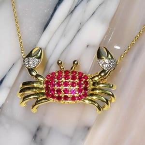 May include: A gold-colored crab pendant necklace. The crab's body is covered in small, red gemstones, with diamond-like accents on the claws. The necklace chain is also gold-colored.